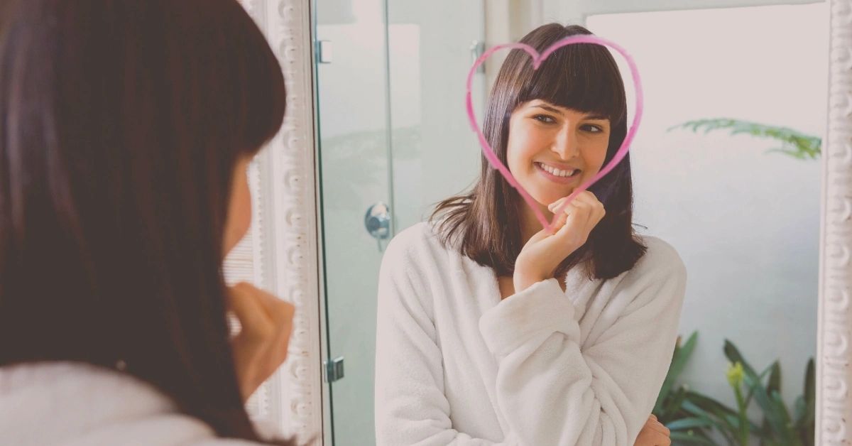 A woman practices positive self-talk in her bathroom mirror.