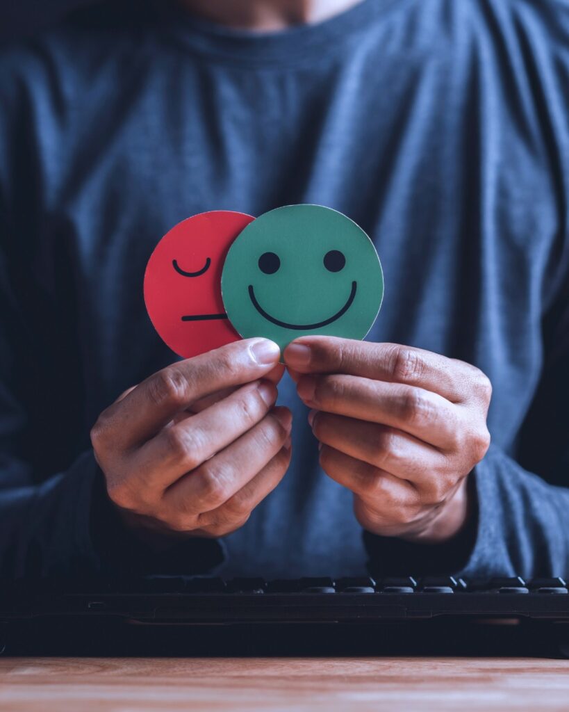 A person in mental distress holds two smiley faces, one happy, one sad, deciding between the two.