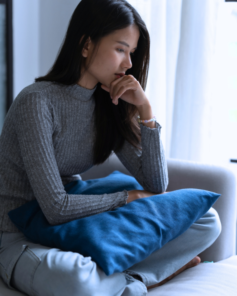 A young asian woman sits cross-legged on a sofa, feeling anxious as she experiences the OCD cycle.