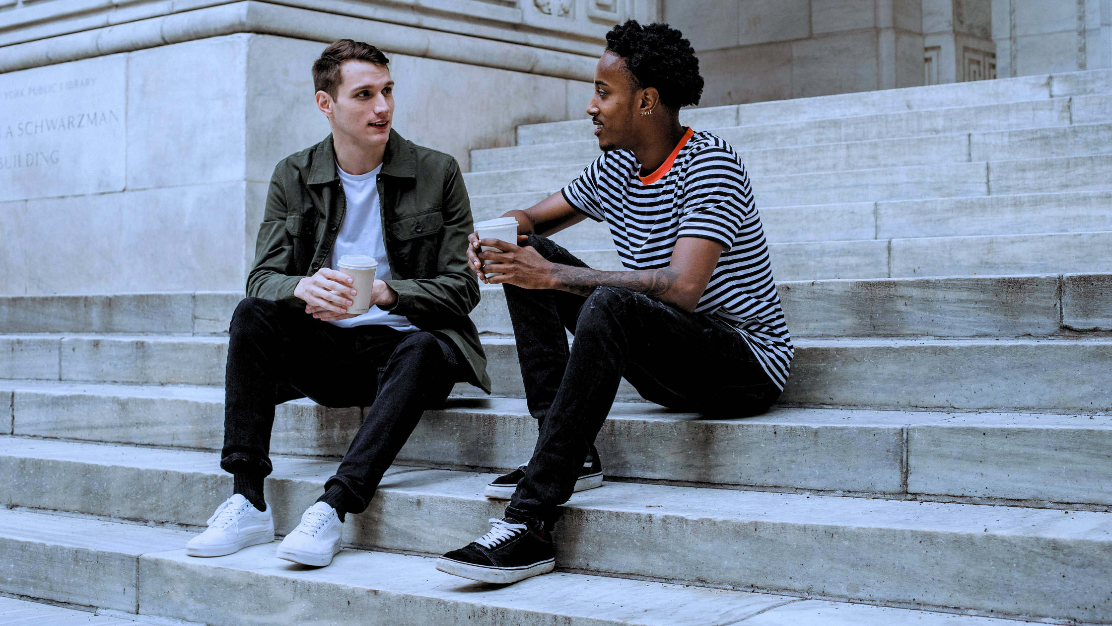 Two met sit on a flight of stairs, talking about mental health, while drinking cups of coffee.